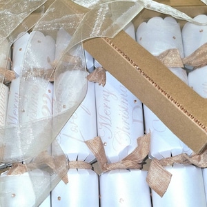 May include: A box of white and gold Christmas crackers with a sheer gold ribbon. The crackers are decorated with the text "Merry Christmas".