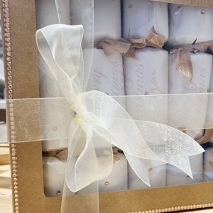 May include: A brown cardboard box with a clear plastic window, filled with white Christmas crackers tied with a beige ribbon. The crackers are decorated with gold glitter and the text "Merry Christmas" is printed on them.