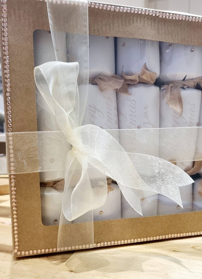 May include: A brown cardboard box with a clear plastic window, filled with white Christmas crackers tied with a beige ribbon. The crackers are decorated with gold glitter and the text "Merry Christmas" is printed on them.