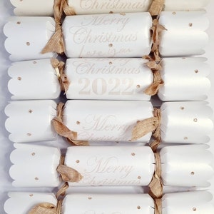 May include: A set of six white Christmas crackers with gold ribbon and gold glitter accents. The crackers have the text "Merry Christmas" and "Christmas 2022" printed on them.