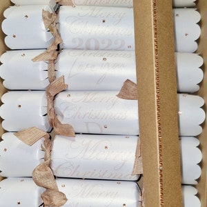 May include: A set of white Christmas crackers with gold ribbon and pink glitter accents. The crackers have the text "Merry Christmas 2022" and "Christmas Love Mom" printed on them.