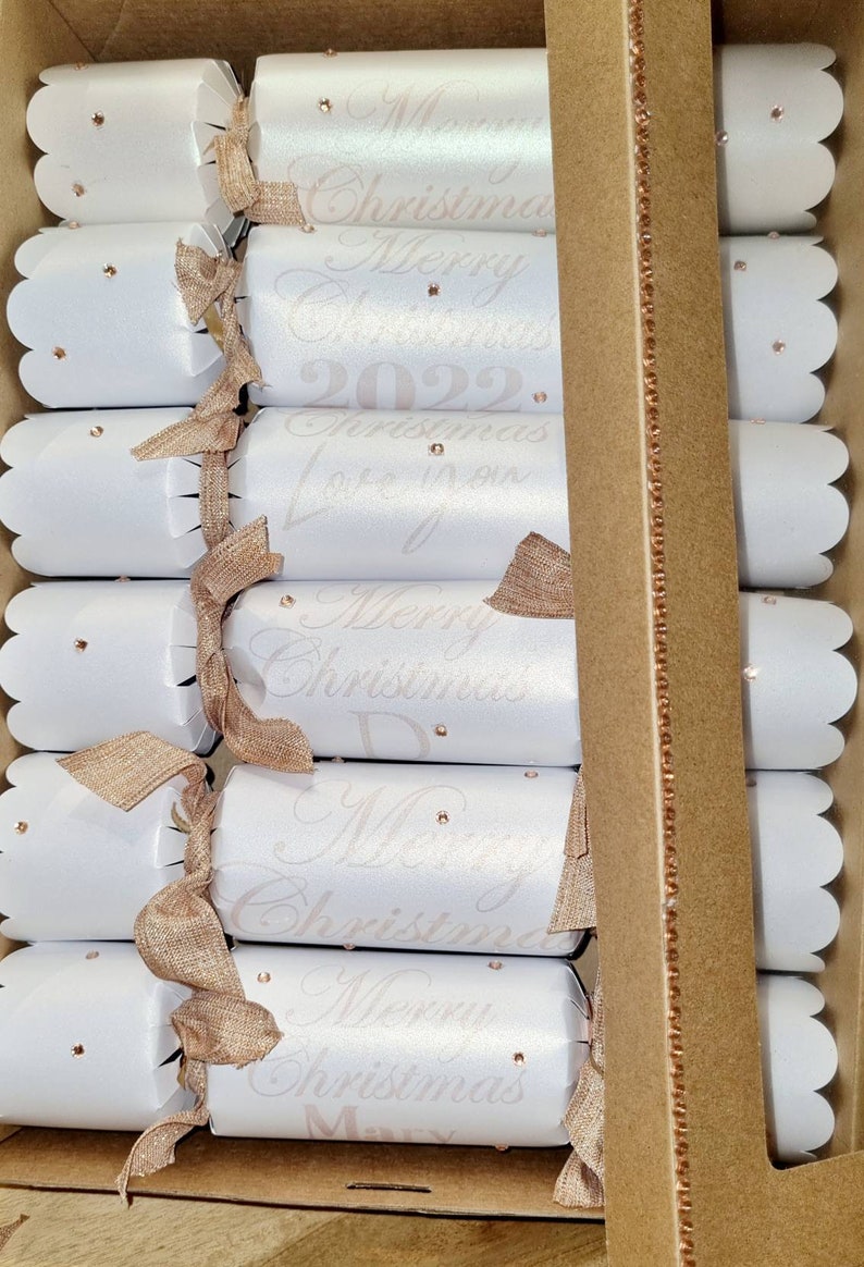 May include: A set of white Christmas crackers with gold ribbon and pink glitter accents. The crackers have the text "Merry Christmas 2022" and "Christmas Love Mom" printed on them.