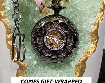 May include: A black and gold pocket watch with a chain, set against a backdrop of green and gold crystals. The watch face is visible, showcasing the intricate inner workings. The text at the bottom reads "COMES GIFT-WRAPPED READY TO TAKE ITS PLACE UNDER THE TREE."