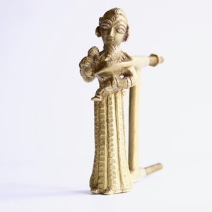 May include: A gold-colored metal figurine of a standing figure with a crown and a beard. The figure is holding a small object in its right hand and has a long, flowing robe. The figurine is attached to a metal rod.