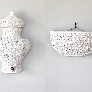 Puede incluir: Dos macetas de pared de cerámica blanca, una con forma de jarrón con tapa y la otra de medio círculo, ambas con un diseño floral.