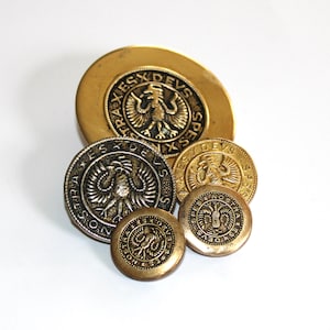 以下が含まれることがあります： 鷲のデザインと「SPES NOSTRA DEVS」という文字が縁に刻まれた、金色の金属製のボタンが5つあります。