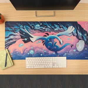 May include: A rectangular desk mat featuring a blue and white creature illustration against a pink and dark blue background with bubbles. The mat is positioned on a wooden desk with a white keyboard, a gray computer mouse, and a green notebook.