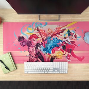 A pink desk mat featuring three video game characters sits on a wooden desk. The characters are all in fighting poses with a dog in the center. A computer monitor, keyboard, and mouse are visible in the background.