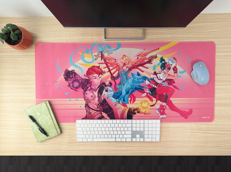 A pink desk mat featuring three video game characters sits on a wooden desk. The characters are all in fighting poses with a dog in the center. A computer monitor, keyboard, and mouse are visible in the background.