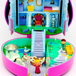 Può includere: Un castello di giocattolo rosa a forma di cuore in plastica con un interno blu e bianco. Il castello ha una scala, un trono, un letto e una cucina. Il castello è aperto e ci sono diverse piccole figure di plastica all'interno, tra cui una principessa, un cavaliere e un cigno.