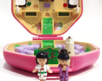 Puede incluir: Una casa de muñecas de plástico rosa con un tobogán verde y dos figuras de pie delante. La casa de muñecas está abierta y el interior es visible. El interior está decorado con muebles y accesorios. Las figuras llevan ropa tradicional.