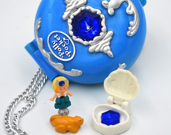 May include: A blue plastic Polly Pocket toy with a silver chain and a blue gem. The toy is open and shows a small blue gem inside. A small plastic doll and a small plastic animal are also included.
