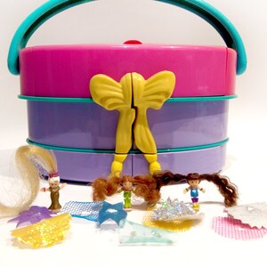 Peut inclure: Une boîte de transport en plastique rose, violette et turquoise avec un nœud jaune. La boîte a une poignée et s'ouvre pour révéler trois compartiments. Il y a quatre petites poupées et des accessoires à l'intérieur de la boîte.