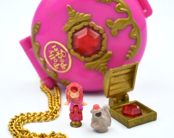 Puede incluir: Un compacto Polly Pocket rosa con una cadena dorada, una gema roja y detalles dorados. El compacto está abierto, revelando una pequeña muñeca y una figurita de ratón.