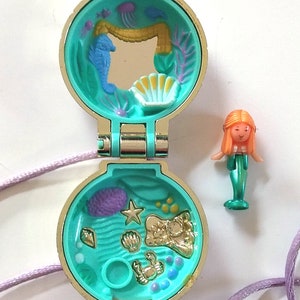Könnte beinhalten: Eine goldfarbene Halskette mit einem türkisfarbenen Anhänger, der sich öffnet und eine Meerjungfrauen-Szene enthüllt. Der Anhänger hat einen Spiegel im Inneren und eine Vielzahl von Meeres-Anhängern. Die Halskette wird mit einer separaten Meerjungfrauen-Figur geliefert.