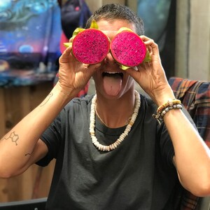 以下が含まれることがあります： ピンクのドラゴンフルーツの半分を2つ、目の前に持って、舌を出している人がいます。黒いTシャツと白いビーズネックレスをしています。