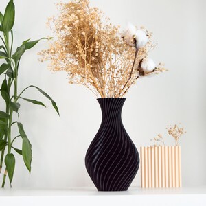 Pu&ograve; includere: Un vaso nero con un design ondulato contiene fiori secchi e boccioli di cotone. Un vaso di legno pi&ugrave; piccolo con scanalature verticali si trova su una superficie bianca.