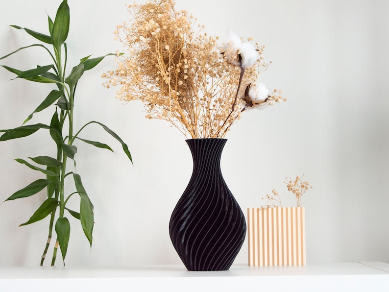 Pu&ograve; includere: Un vaso nero con un design ondulato contiene fiori secchi e boccioli di cotone. Un vaso di legno pi&ugrave; piccolo con scanalature verticali si trova su una superficie bianca.