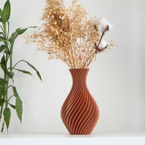 Pu&ograve; includere: Un vaso di legno marrone con un design a spirale, che contiene fiori secchi e boccioli di cotone. Il vaso &egrave; su una superficie bianca.