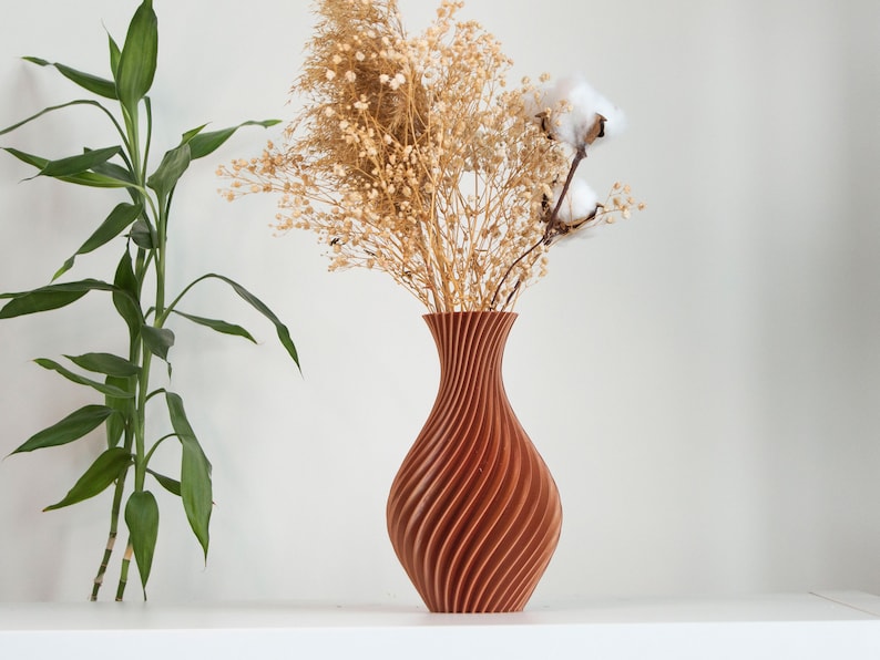 Pu&ograve; includere: Un vaso di legno marrone con un design a spirale, che contiene fiori secchi e boccioli di cotone. Il vaso &egrave; su una superficie bianca.