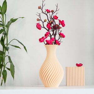 Pu&ograve; includere: Un vaso in ceramica beige chiaro con un design a spirale e texture. Il vaso &egrave; riempito con un bouquet di fiori artificiali rosa con steli marrone scuro. Il vaso &egrave; su una superficie bianca.