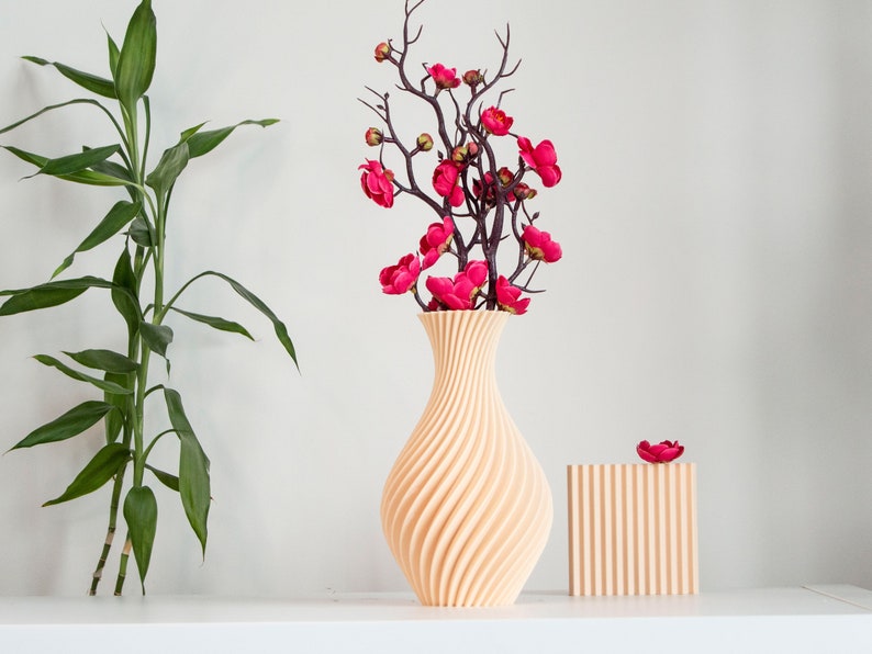 Pu&ograve; includere: Un vaso in ceramica beige chiaro con un design a spirale e texture. Il vaso &egrave; riempito con un bouquet di fiori artificiali rosa con steli marrone scuro. Il vaso &egrave; su una superficie bianca.