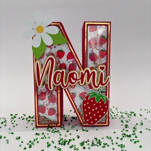 May include: A red and gold letter N with a strawberry and daisy design. The letter is decorated with glitter and has the name "Naomi" written in gold letters on it.