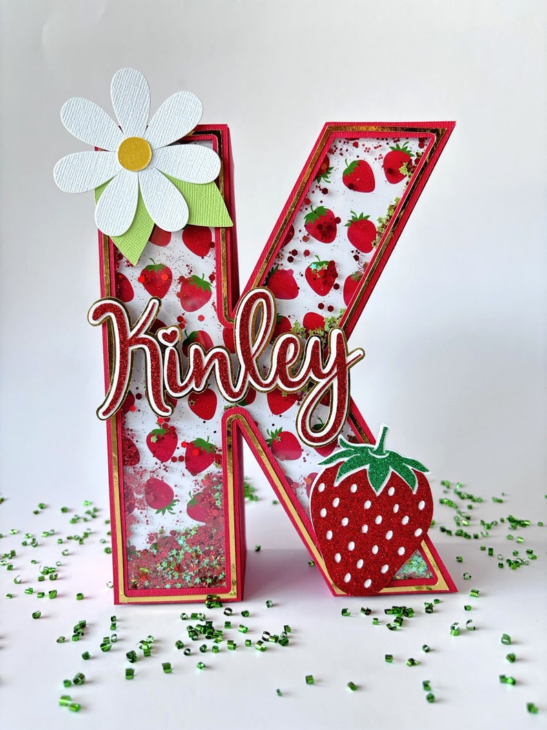 May include: A red and gold letter K with a strawberry pattern and glitter inside. The letter is decorated with a white daisy and a red strawberry. The name "Kinley" is written in white cursive on the letter.