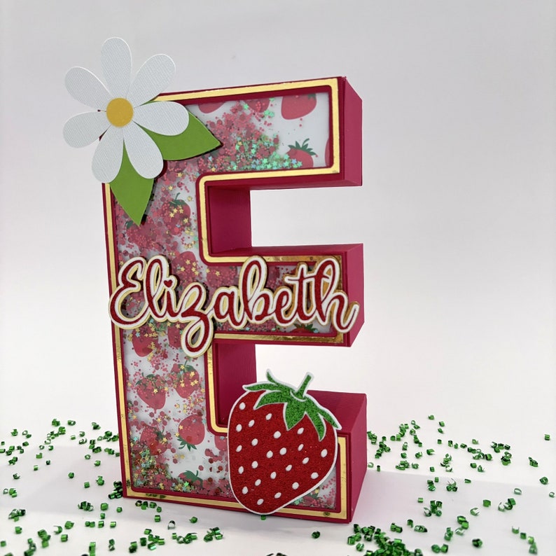 May include: A pink letter E with a gold frame, filled with a strawberry and glitter pattern. The letter has a white daisy with green leaves on top and the name "Elizabeth" in red letters on the front.