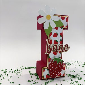 May include: A red and white cardboard letter I decorated with a strawberry pattern, a white daisy, and green leaves. The letter has a gold border and the name "Isaac" is written in red cursive letters.