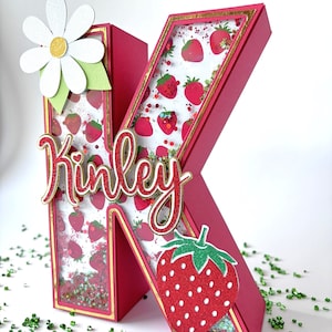 May include: A pink letter K with a gold border and a strawberry pattern inside. The letter is decorated with a white daisy and a red strawberry. The letter is spelled "Kinley".