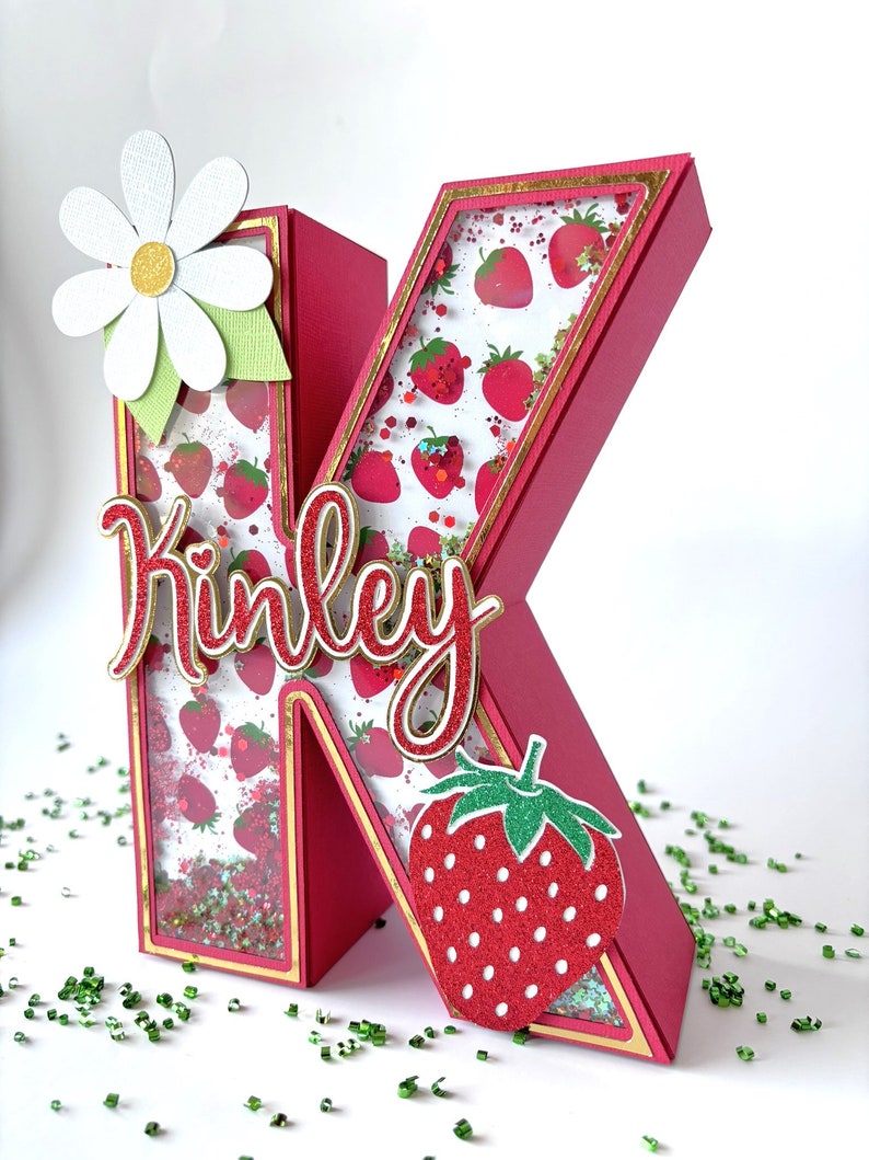 May include: A pink letter K with a gold border and a strawberry pattern inside. The letter is decorated with a white daisy and a red strawberry. The letter is spelled "Kinley".