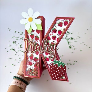 May include: A red, glitter-covered letter K with a white daisy and a strawberry on the side. The letter is decorated with a strawberry pattern and the name "Kinley" is written in gold cursive.