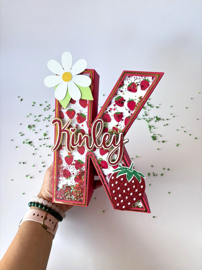 May include: A red, glitter-covered letter K with a white daisy and a strawberry on the side. The letter is decorated with a strawberry pattern and the name "Kinley" is written in gold cursive.