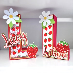 May include: Two red and white cardboard letters, L and I, decorated with strawberries and glitter. The letters are topped with white daisies and have the names "Lucy" and "Leilani" written in red glitter.
