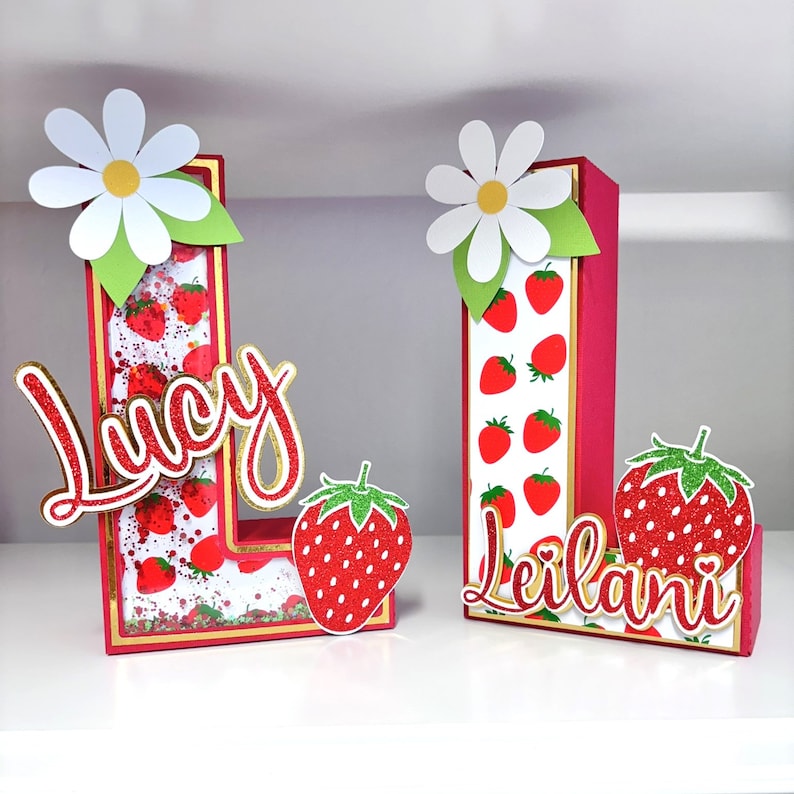 May include: Two red and white cardboard letters, L and I, decorated with strawberries and glitter. The letters are topped with white daisies and have the names "Lucy" and "Leilani" written in red glitter.
