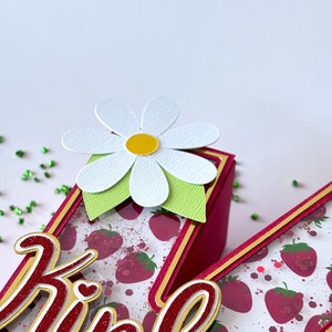 May include: A red and gold letter "K" with a white daisy on top. The letter is decorated with a strawberry pattern and glitter. The name "Kinley" is written in red and gold glitter on the letter.