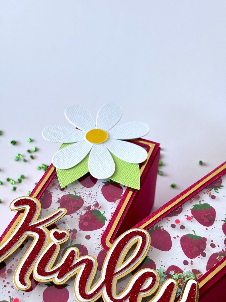 May include: A red and gold letter "K" with a white daisy on top. The letter is decorated with a strawberry pattern and glitter. The name "Kinley" is written in red and gold glitter on the letter.
