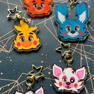 May include: Five acrylic keychains featuring cartoon character heads with gold-coloured star-shaped clasps. The characters include an orange bear with a top hat, a blue rabbit, a yellow bird, and two white foxes with pink accents and red cheeks.
