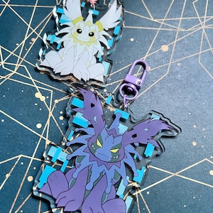 May include: Two acrylic keyrings featuring cartoon monster designs. One is white with pink hardware, the other is purple and blue with purple hardware. Both have a clear border and a blue geometric design.