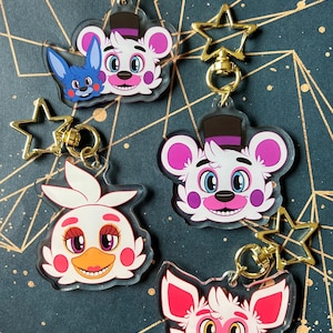 May include: Four acrylic keychains featuring cartoon character heads with gold-coloured star-shaped clasps. The designs include a white bear with a purple hat, a blue rabbit, a white bird with a yellow beak, and a white fox with pink accents.