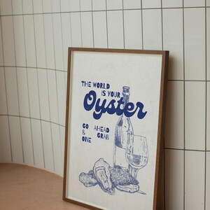 May include: Framed print with a blue illustration of oysters, a wine bottle, and a glass. The text "THE WORLD IS YOUR OYSTER" is displayed in blue, along with "GO AHEAD & GRAB ONE". The frame is brown.