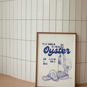 May include: A framed print with a vintage-style illustration in blue. The artwork features a wine bottle, a wine glass, and oysters, with the text "THE WORLD IS YOUR OYSTER." The phrase "GO AHEAD & GRAB ONE" is also included.
