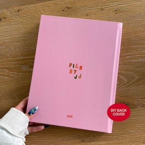 Pu&ograve; includere: Un libro con copertina rigida rosa chiaro con il testo FILM BY JJ stampato in piccole lettere maiuscole multicolori al centro e l'anno 2025 stampato in piccolo testo arancione vicino al bordo inferiore della copertina.