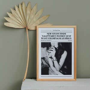 May include: A framed black and white photo of a woman holding a glass of champagne. The photo has a newspaper headline that reads "NEW STUDY FINDS 'SAGITTARIUS WOMEN' JUST WANT CHAMPAGNE & DISCO" with the date "November 22 - December 21" and the text "The Archer".