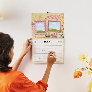 May include: A decorative wall calendar for May 2026, featuring two photo frames and floral accents. The calendar is being marked with a pencil. A yellow and white striped vase holds a bouquet of colorful flowers.