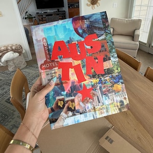 Op de afbeelding: Een hardcover boek met de titel AUSTIN in grote rode letters en een ster op de kaft. De kaft bevat een collage van stadsgezichten, waaronder een motelbord, een skyline en twee handen die drankjes vasthouden met labels in de vorm van Texas.