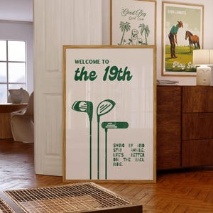 May include: A framed print with the text "WELCOME TO the 19th" and golf club illustrations in green. Two other framed prints are visible, one with a dog and palm trees, and another with a cowboy and horse. The room has wooden floors.