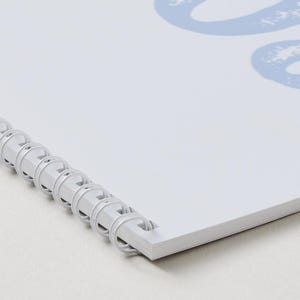 May include: A white spiral-bound notebook featuring a light blue graphic design on the cover. The binding consists of a white plastic coil that holds the pages together along the left edge. The cover displays a partial light blue number 202.