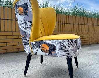 May include: An accent chair featuring a mustard yellow velvet interior with vertical channel tufting and a back and base upholstered in a gray and white fabric with a black, white, and orange toucan and tropical leaf print on four black legs.
